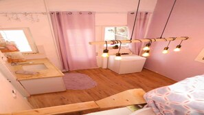 A small, cozy bedroom with a wooden bed, a white bedside table, and a pink wall. There is a window with a curtain, a small white cabinet, and a wooden stool.