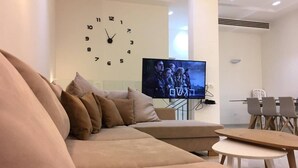 Ein modernes Wohnzimmer mit einem beigen Sofa, einem Flachbildfernseher, einer Wanduhr und einem Essbereich mit Stühlen und einem Tisch.