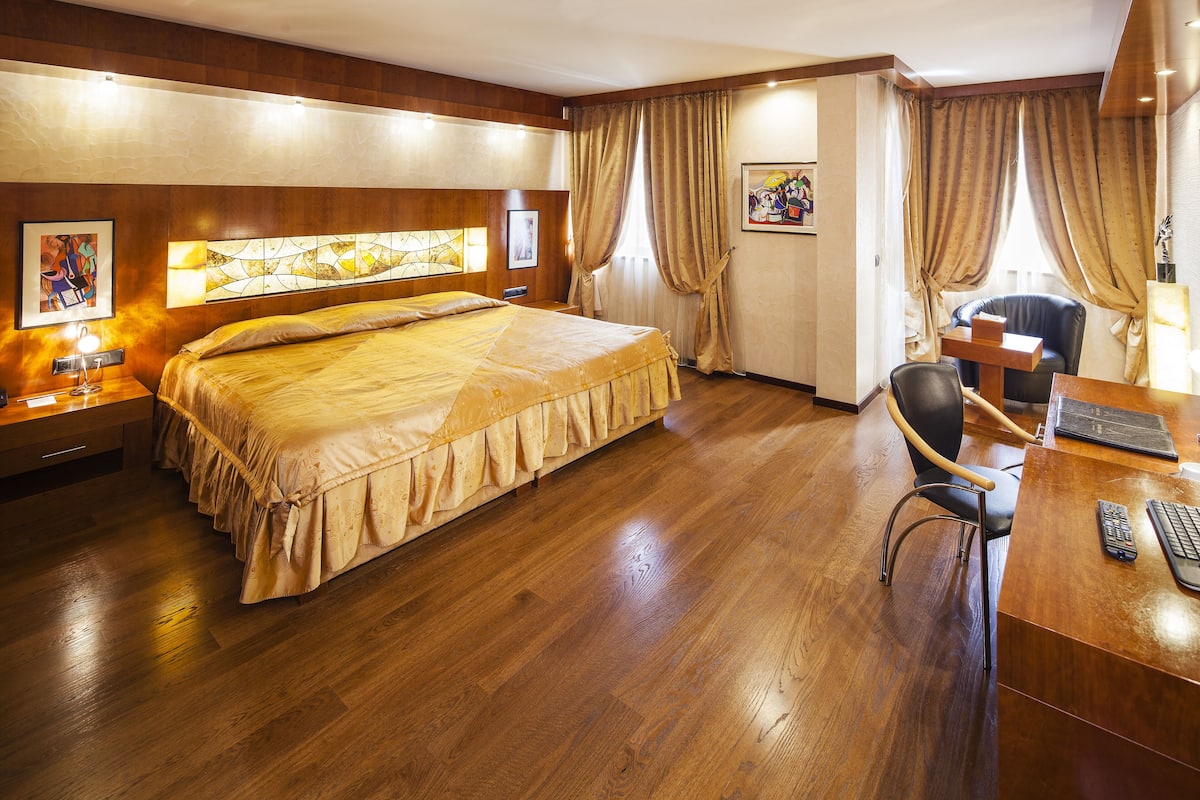 Een hotelkamer met een groot bed, houten vloeren, een bureau met stoel en een raam met gordijnen.