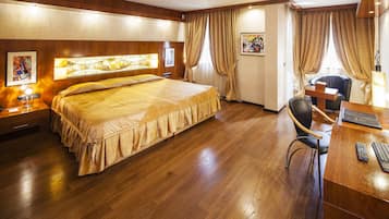 A hotel room with a large bed, wooden flooring, a desk with a chair, and a window with curtains.