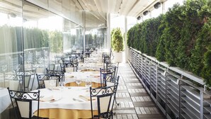Un espace extérieur abrité, aménagé pour les repas, avec des tables pour deux personnes, entouré de murs de verre et d’une rambarde en métal.