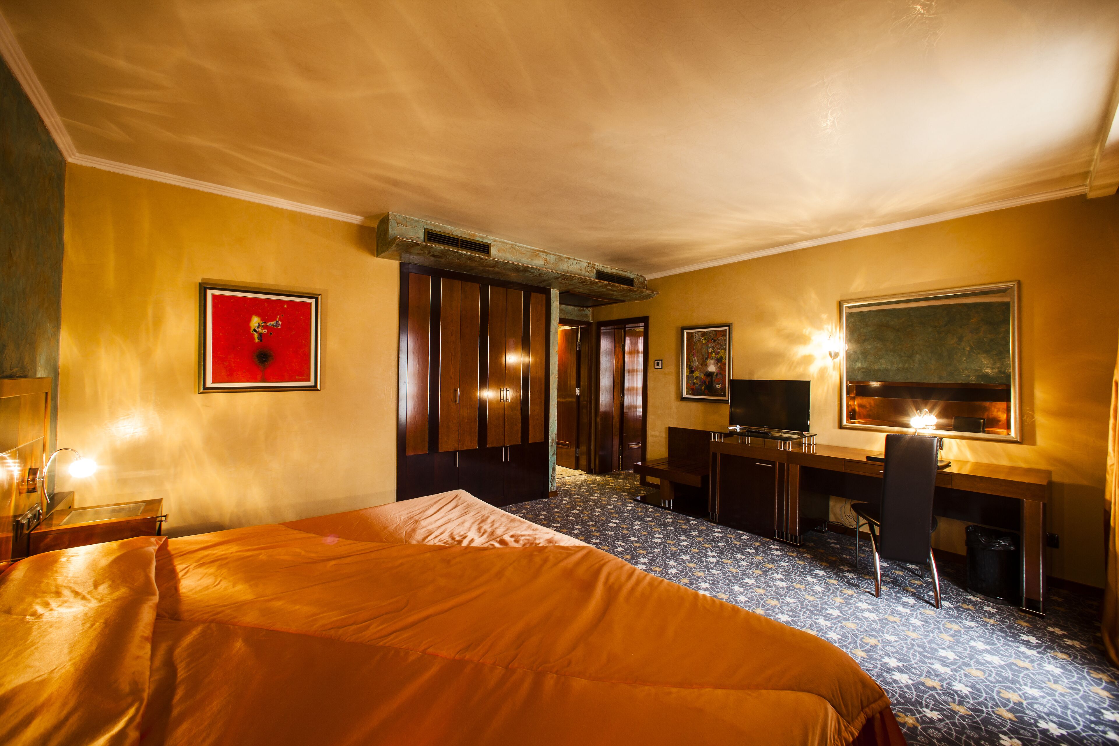 A hotel room with a bed, desk, chair, television, and artwork on the walls.