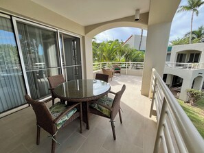 A balcony with a glass table and wicker chairs, overlooking a lush garden and a building with a white exterior.