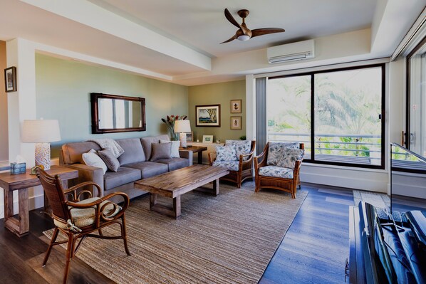 A spacious living room with a sofa, chairs, a coffee table, and a piano. There is a large window with a view of greenery outside.