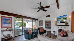 Un salon spacieux doté d’une grande fenêtre, d’un ventilateur de plafond et d’une piscine visible à travers les portes vitrées.