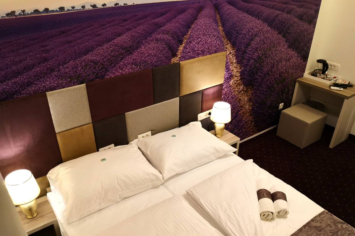 A hotel room with a large bed, two bedside lamps, a nightstand, a small table, and a wall tapestry depicting a lavender field.