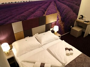 Una habitació d'hotel amb un llit gran, dues làmpades de nit, una tauleta de nit, una taula petita i un teixit mural que representa un camp de lavanda.