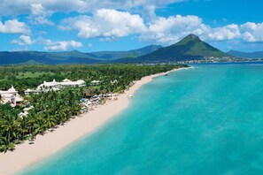 Un resort costero con una playa de arena, palmeras y una montaña al fondo.