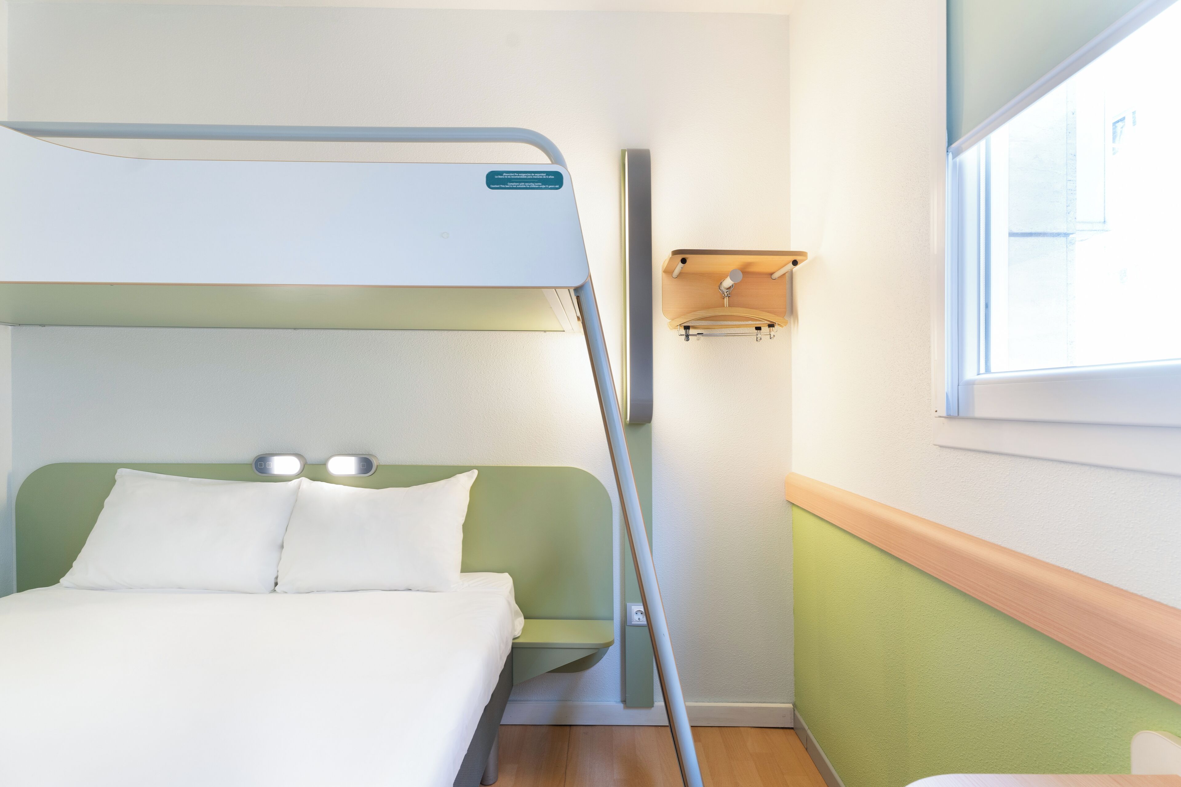 Una cama litera con cabecera verde y ropa de cama blanca, un pequeño estante de madera con ganchos y una ventana con vistas al exterior.