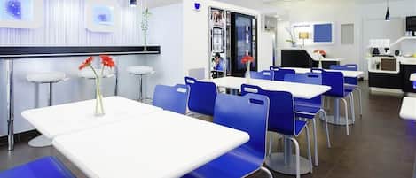 A modern dining area with white tables and blue chairs, a reception desk, and a counter with a coffee machine.