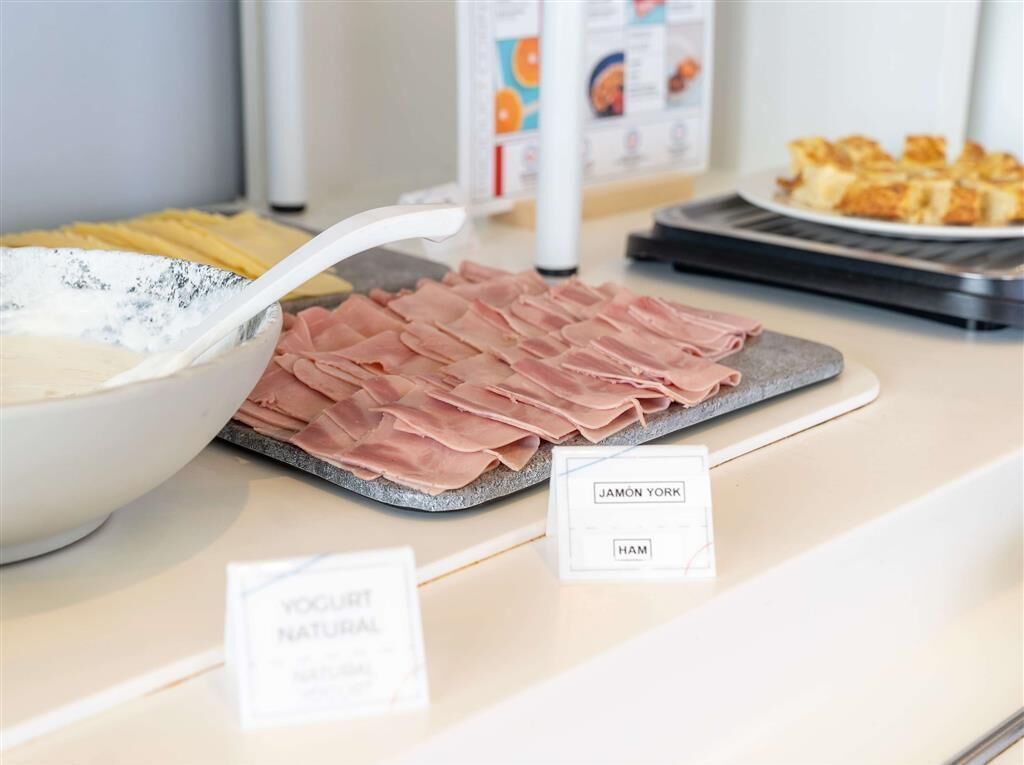 A buffet counter with a variety of food items including a bowl of yogurt, sliced ham, and a plate of pastries.