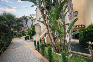 Un chemin pavé mène à un bâtiment doté d’un escalier, le tout étant entouré d’une végétation luxuriante et surplombé par un ciel d égagé.