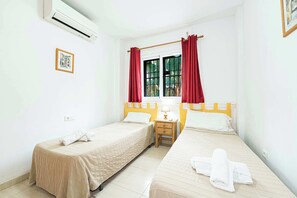 Ein Hotelzimmer mit zwei Betten, einem Fenster mit roten Vorhängen, einem Nachttisch mit Lampe und einer Klimaanlage.