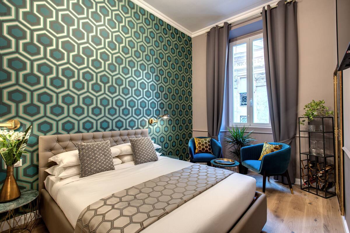A modern hotel room with a large bed, a blue armchair, a small round table, and a bookshelf.