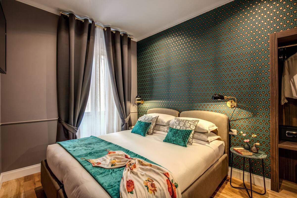 A neatly arranged bedroom with a bed, curtains, a bedside table with a lamp, and a patterned wall.