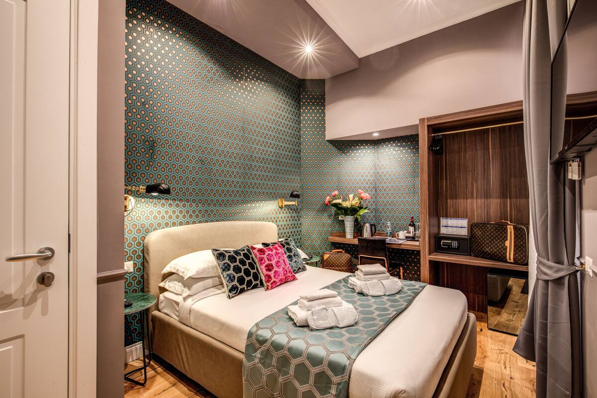 A hotel room with a patterned wall, a bed with pillows, a bedside table with a radio, and a wardrobe.