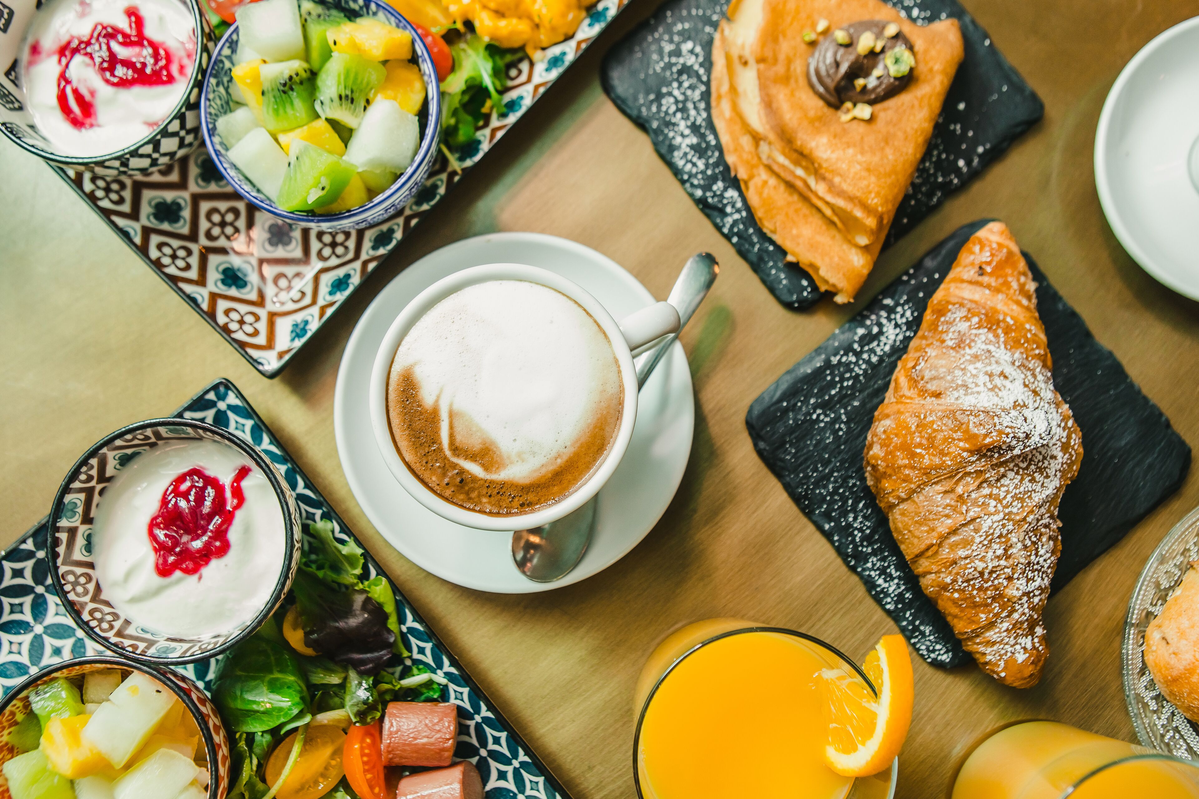 En frukostuppdukning med en kopp kaffe, croissanter, frukt och ett glas apelsinjuice på ett träbord.