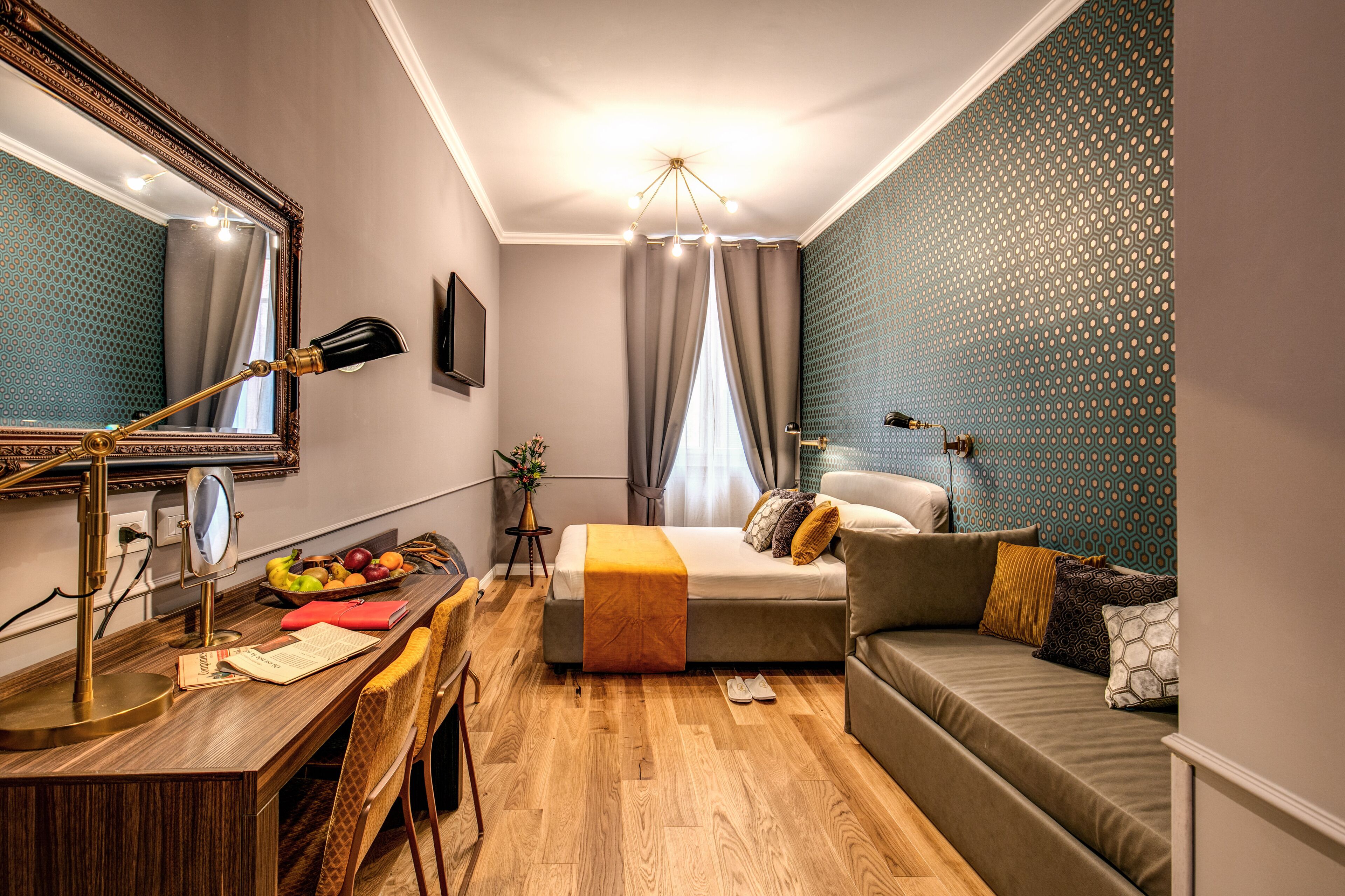 A modern hotel room with a wooden desk, a bed with a beige blanket, a green sofa, and a patterned wall.