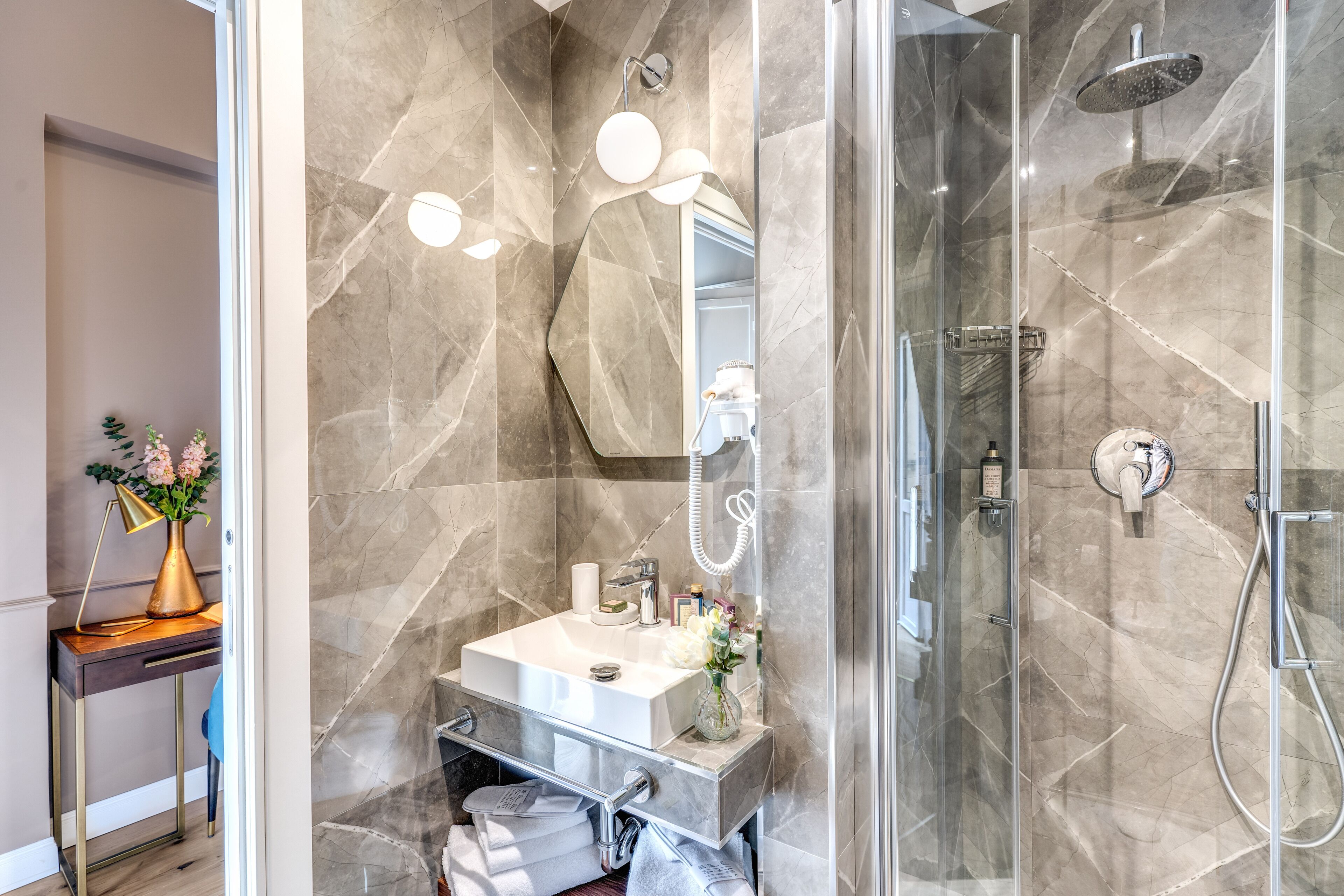A modern bathroom with a glass-enclosed shower, a marble vanity, and a small desk with a vase of flowers.