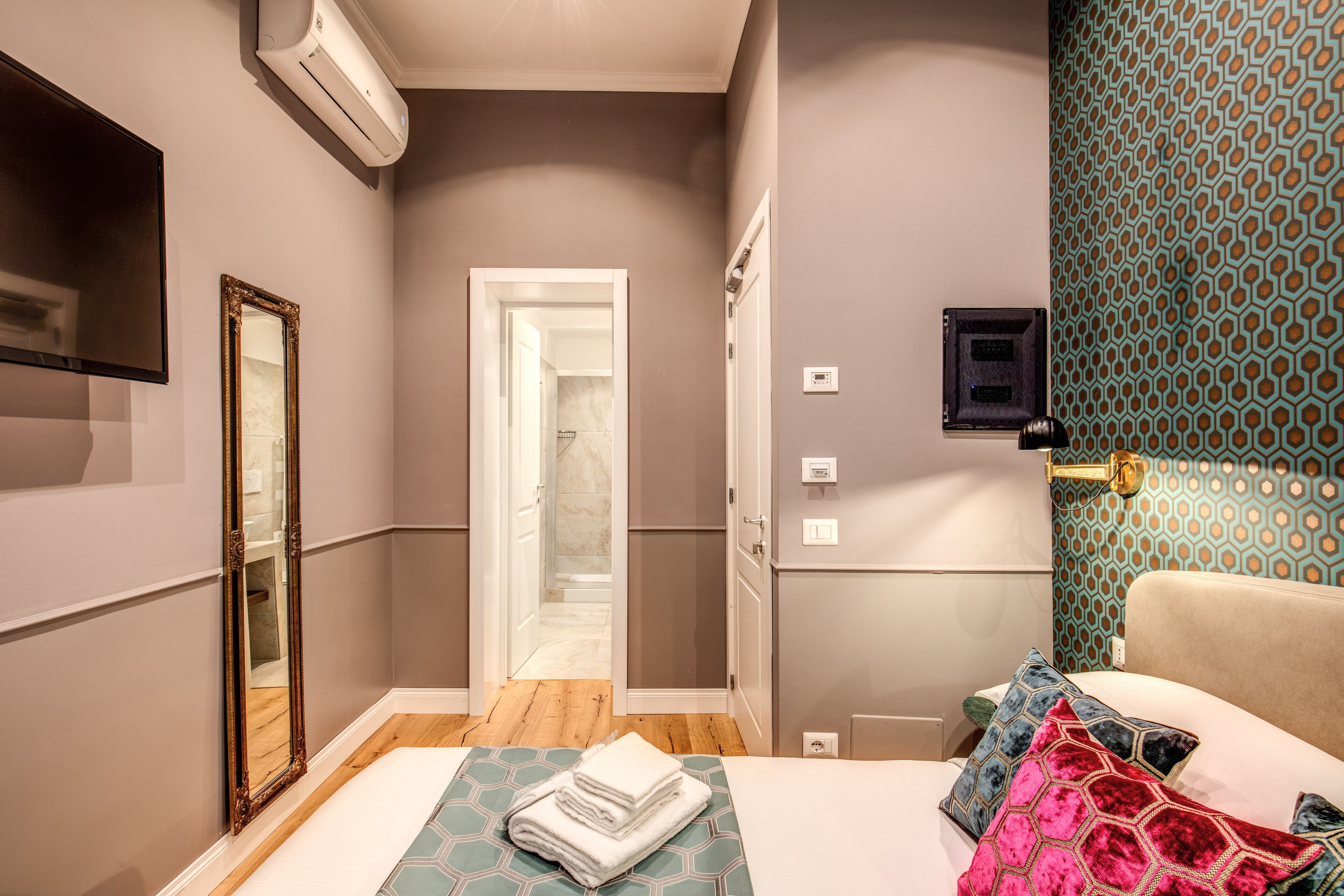 A hotel room with a bed, a mirror, an air conditioning unit, a door leading to a bathroom, and a patterned wall.