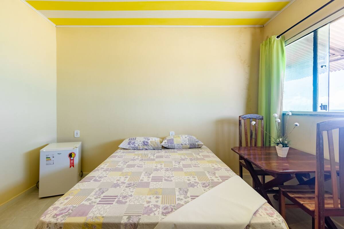 A small room with a bed, a table, a chair, a refrigerator, and a window with curtains.
