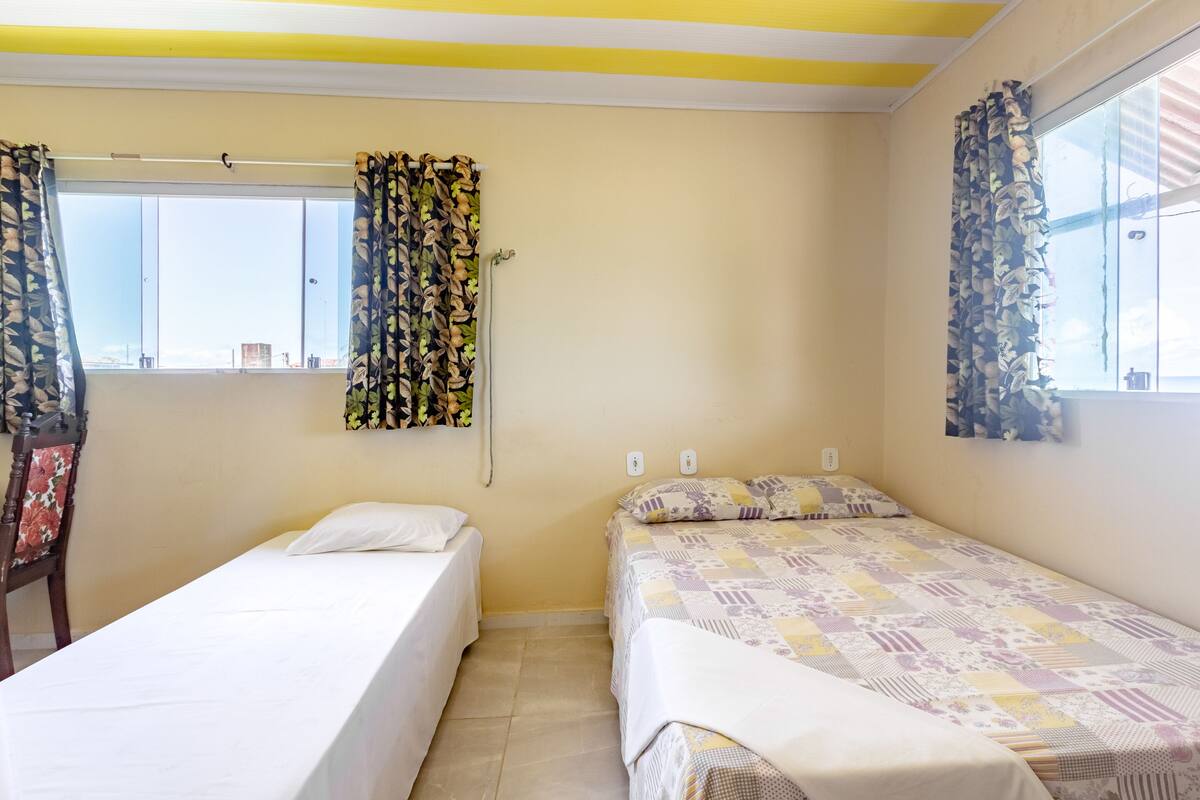 A room with two single beds, a window with curtains, a chair, and a wall-mounted light fixture.