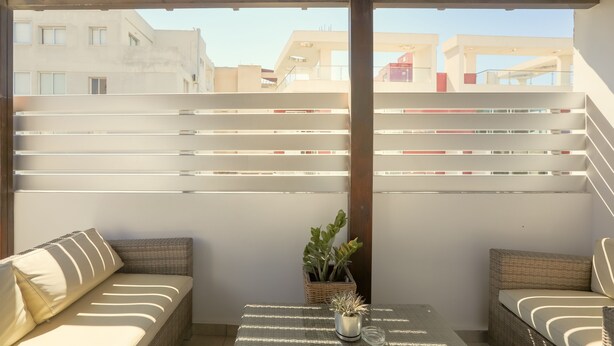 A covered outdoor seating area with wicker furniture and a small table, featuring a view of buildings in the background. - Yiolanda - 2BR - City Center (Larnaca)