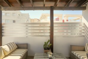 Un espacio al aire libre con techo, amueblado con sillas de mimbre y una pequeña mesa, que ofrece vistas a edificios en el fondo.