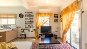 A modern living room with a flat-screen TV, a coffee table, and a sofa. There is a kitchen area with a sink and cabinets, and a large window with curtains. - Yiolanda - 2BR - City Center (Larnaca)