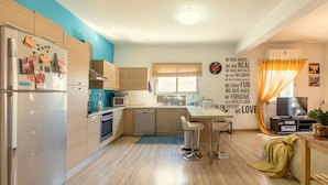 A modern kitchen with wooden cabinets, a built-in microwave, and a refrigerator. There is a dining area with a table and chairs, and a window with curtains. - Yiolanda - 2BR - City Center (Larnaca)