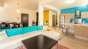 A modern living room with a sofa, coffee table, and a dining area with chairs and a table. - Yiolanda - 2BR - City Center (Larnaca)