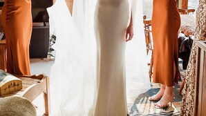 Una mujer ajusta un vestido de novia en un espacio interior con paredes de madera y ventanas amplias.