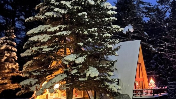 Una cabaña cubierta de nieve con luz cálida en su interior, rodeada de árboles cargados de nieve.