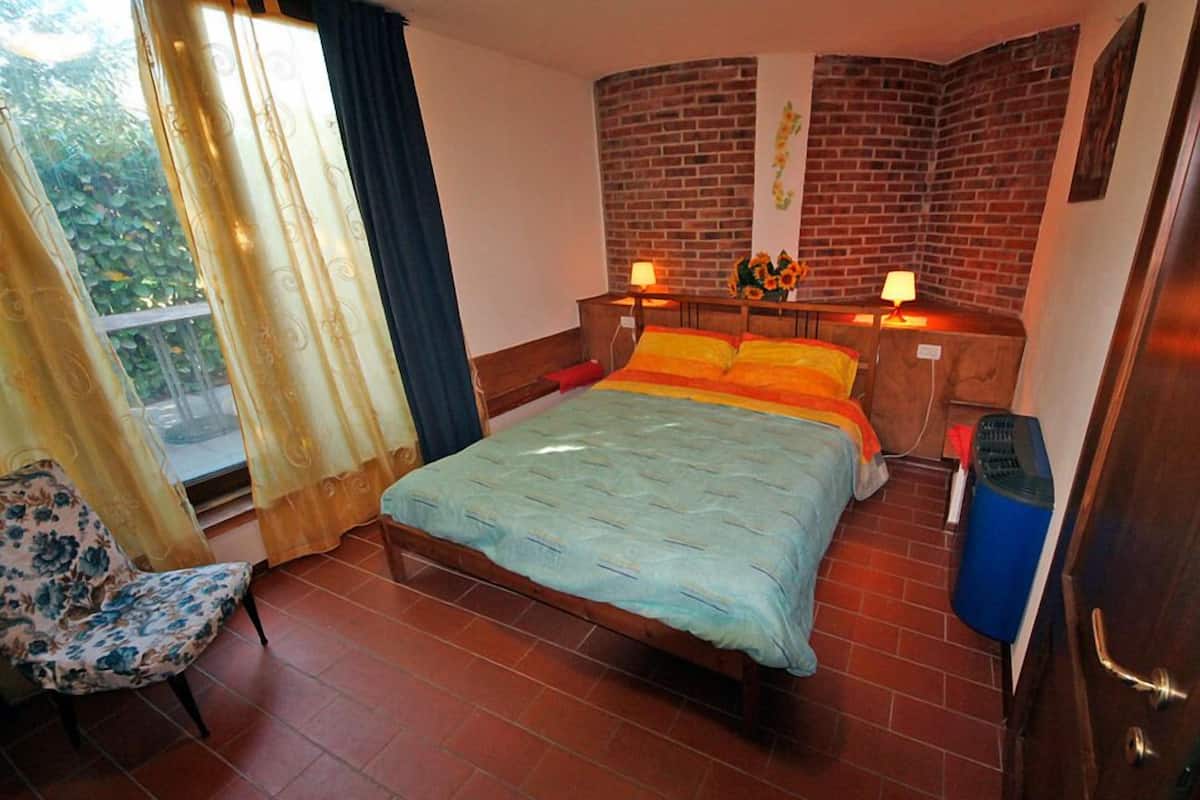 Ein Schlafzimmer mit einem Bett, zwei Nachttischlampen, einem Sessel, einem Fenster mit Vorhängen und einer Ziegelwand.
