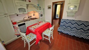 Una cocina compacta con armarios blancos, una mesa de comedor con mantel rojo, dos sillas y un sofá estampado.