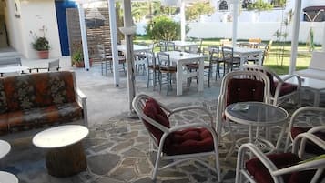 Eine Terrasse im Freien mit Steinböden, weißen Tischen und Stühlen sowie einer Couch mit roten Kissen.
