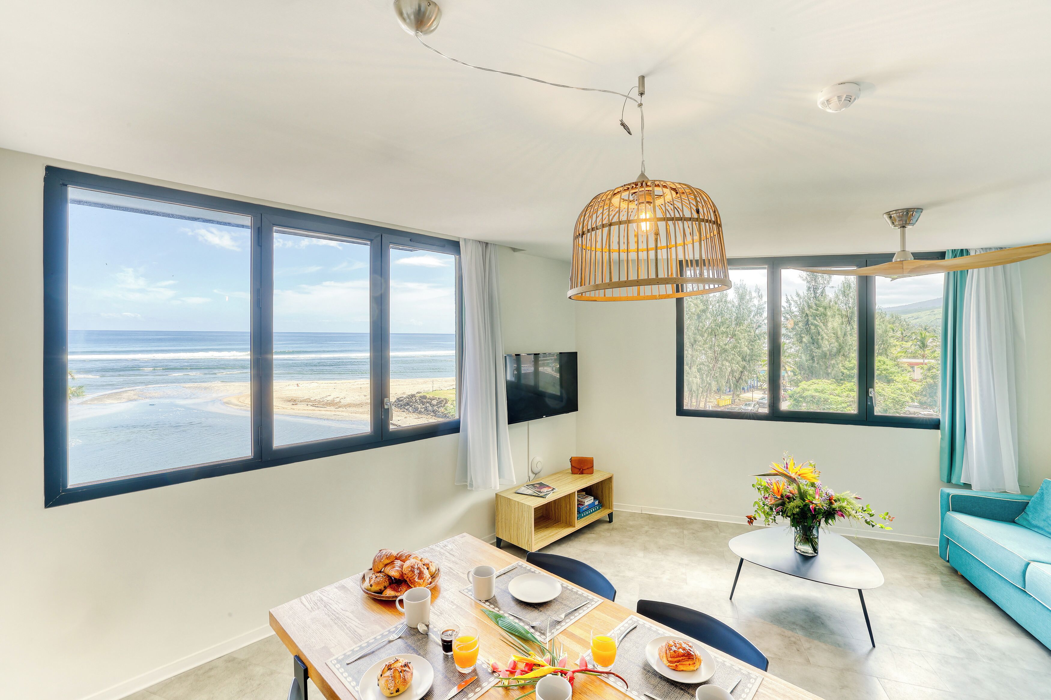Une chambre moderne avec de grandes fenêtres, une table à manger dressée pour le petit-déjeuner, un canapé bleu et un téléviseur.
