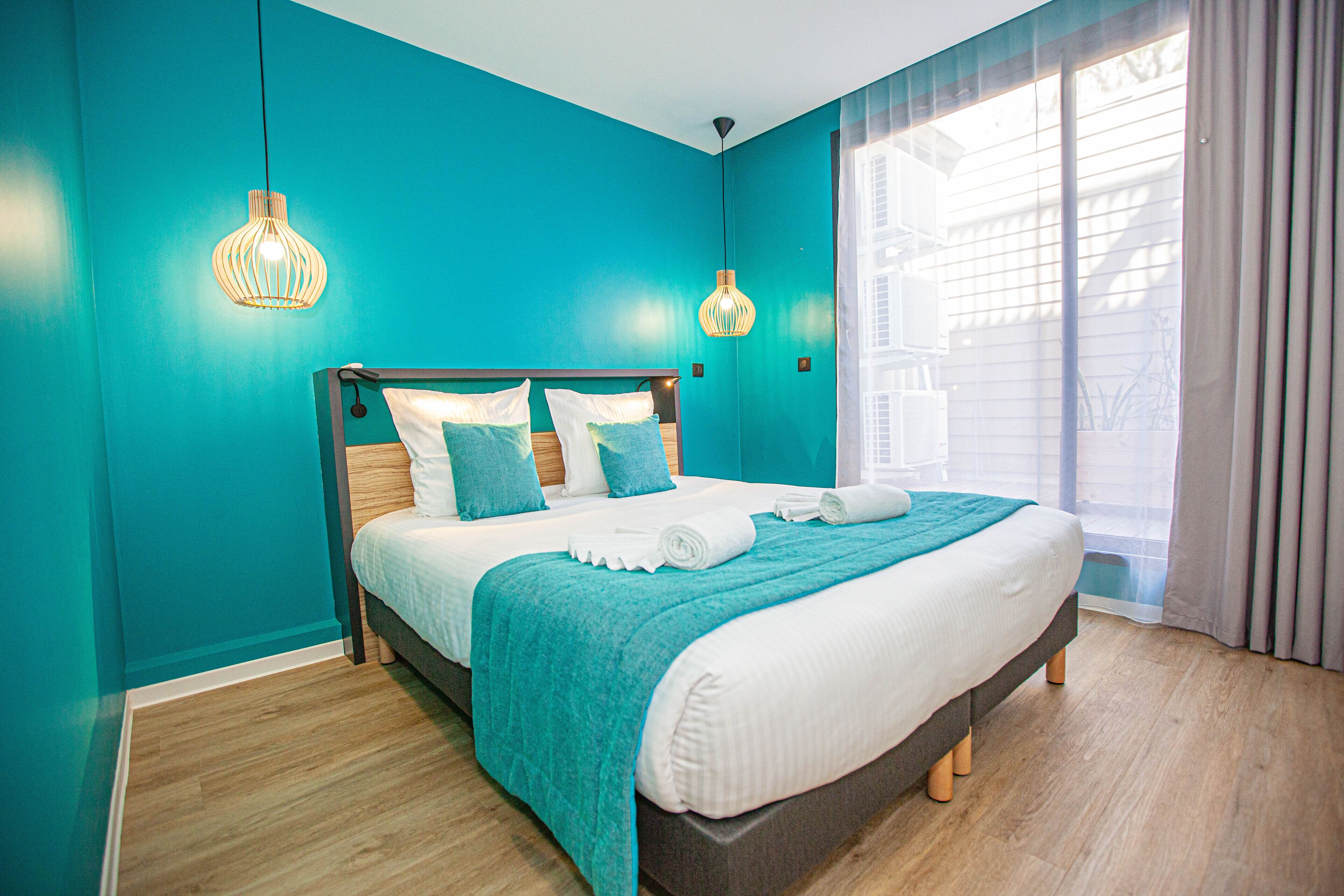 A bedroom with teal walls, a wooden bed frame, white bedding, and two pendant lights.