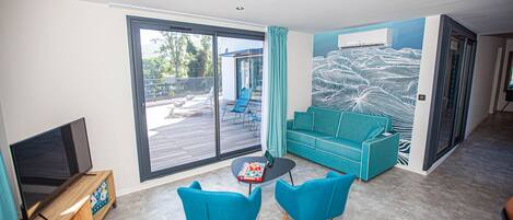 A modern living room with a large sliding glass door to the outdoors, a teal sofa, two matching chairs, a small coffee table, and a flat-screen TV on a wooden stand.