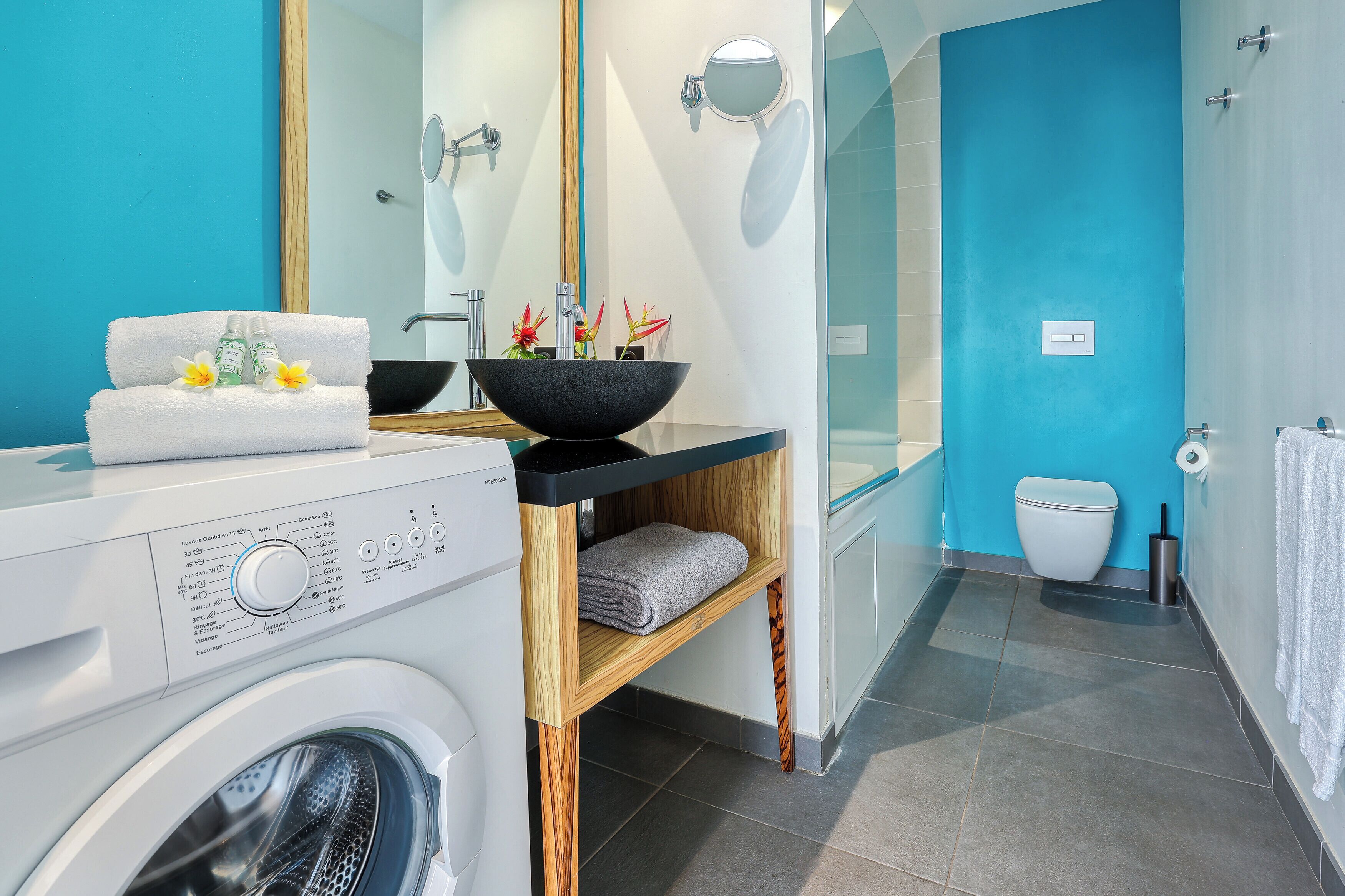 Ein modernes Badezimmer mit Waschmaschine, Waschbecken mit schwarzer Schüssel, Handtuchhalter und einer Duschkabine aus Glas.
