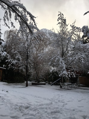 A snow-covered backyard with trees, a swing set, and a wooden deck. - El Encanto Del Manzano - Hostel (San José de Maipo)