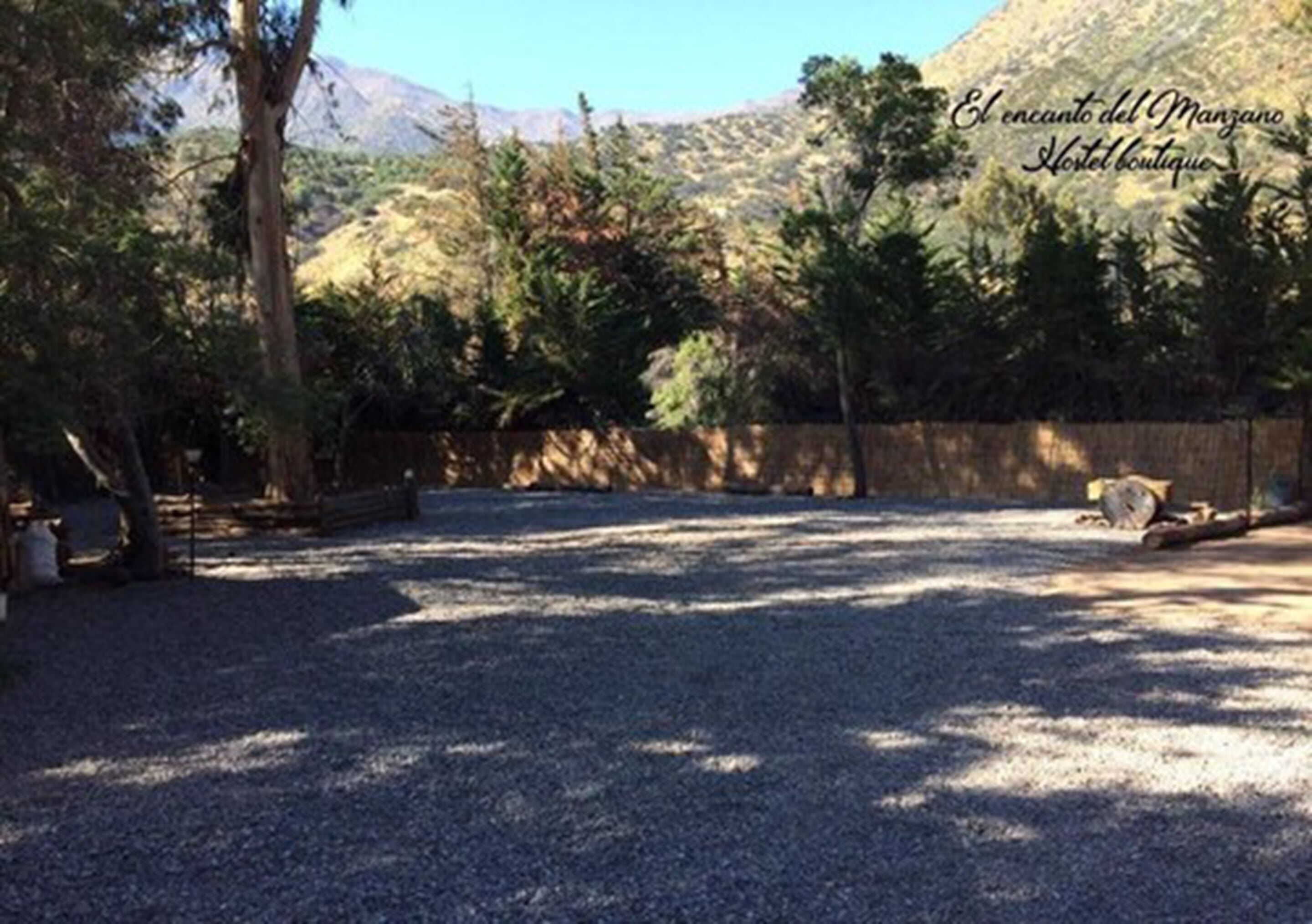 Um estacionamento de cascalho com árvores e um muro de pedra, emoldurado por uma paisagem montanhosa.