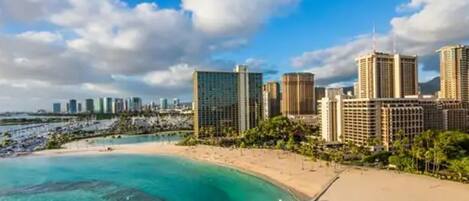 A coastal cityscape with high-rise buildings, a sandy beach, and a pier extending into the turquoise waters.