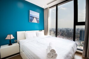 A hotel room with a large bed, a nightstand with a lamp, a city view through large windows, and a picture on the wall.