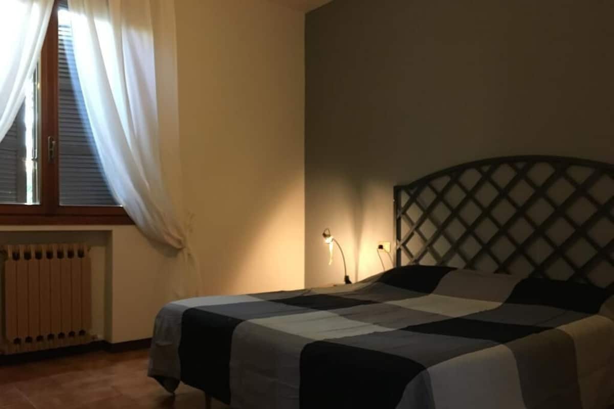 A bedroom with a bed, a radiator, a window with curtains, and a wall-mounted lamp.