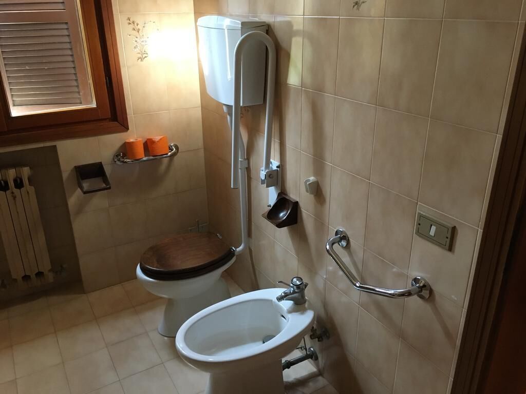 A bathroom with a toilet, bidet, and sink. There is a handrail on the wall and a window with blinds.