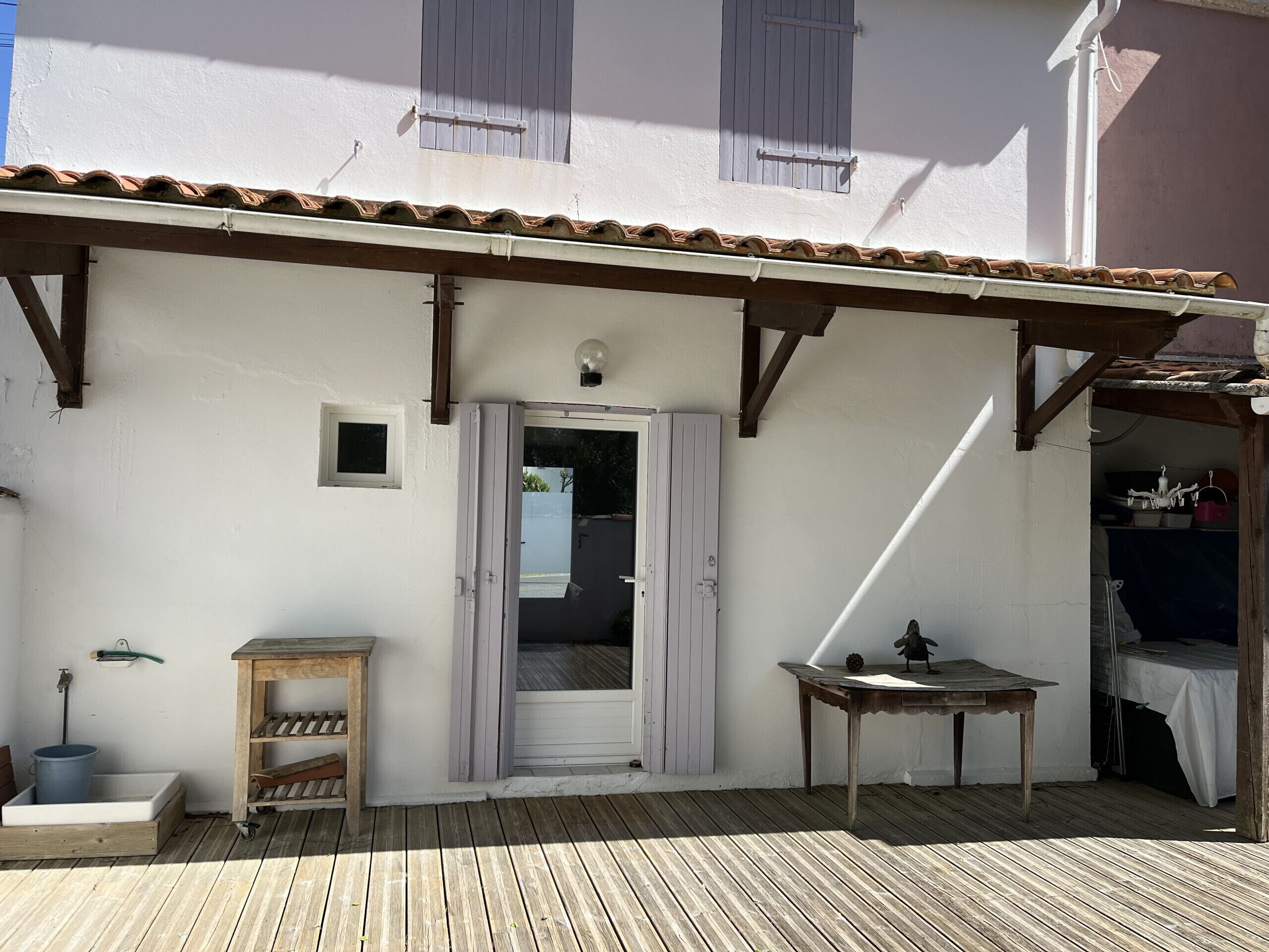 A wooden deck with a white building, a door, a small table, and a shelf with a bucket.