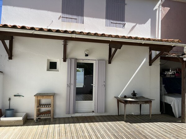 Eine Holzterrasse mit einem weißen Gebäude, einer Tür, einem kleinen Tisch und einem Regal mit einem Eimer.