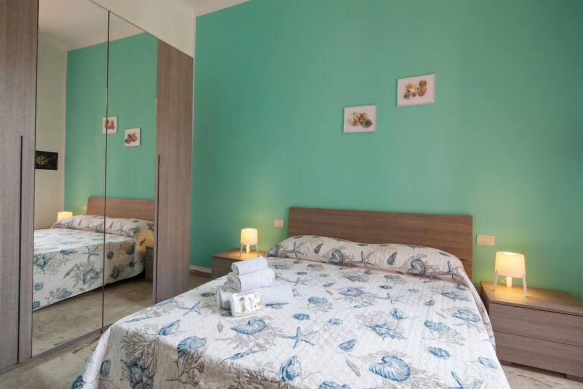 Een slaapkamer met een spiegelkast, een bed met bloemenbedekking, twee nachtkastjes met lampen en een groene muur met ingelijste foto’s.