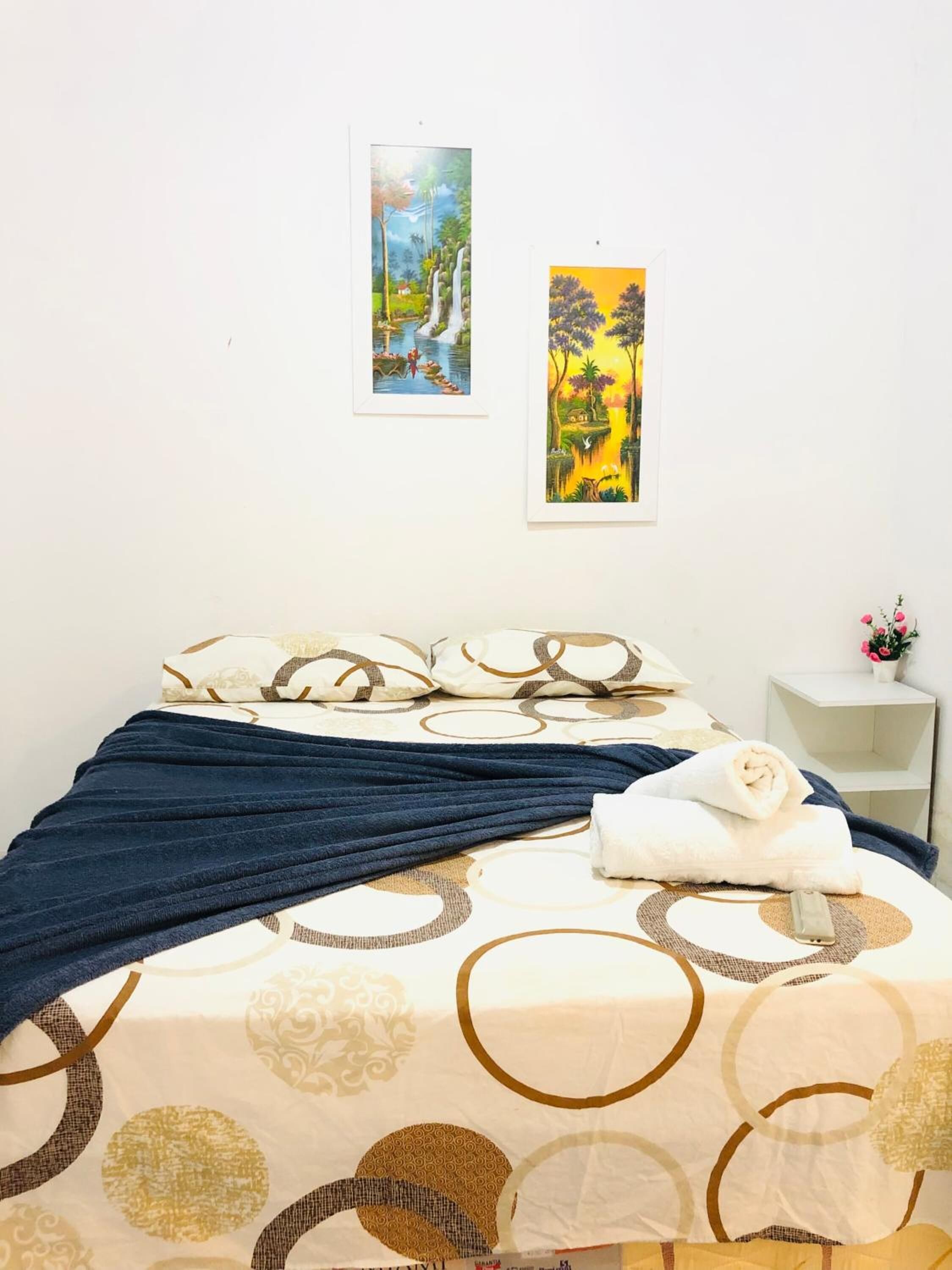 A neatly made bed with a patterned bedspread, a blue blanket, and a rolled white towel. There are two framed paintings on the wall, one depicting a landscape and the other tropical scenery. A small white nightstand with a vase of flowers is visible to the right.